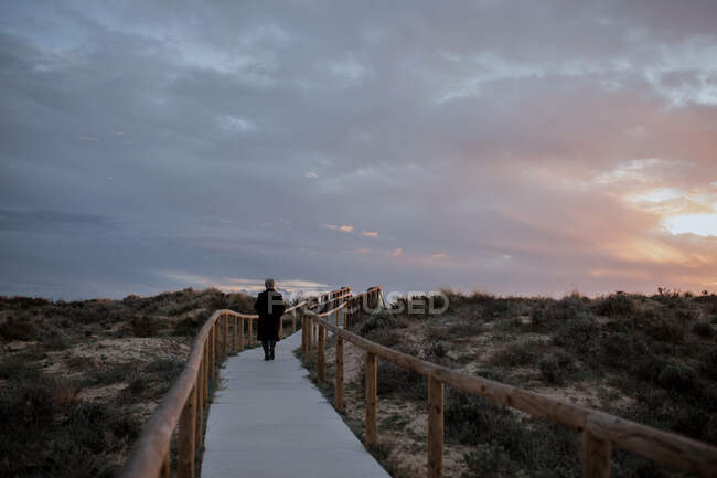 Vue arrière de voyageuse âgée anonyme en vêtements décontractés marchant sur une jetée en bois sur une plage de sable et profitant du paysage marin au coucher du soleil — Photo de stock