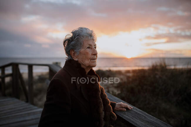 Vista lateral de una anciana viajera vestida con ropa casual parada en un muelle de madera en la playa de arena y disfrutando del paisaje marino al atardecer - foto de stock