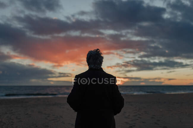 Вид ззаду на невпізнавану старшу жінку в теплому повсякденному одязі, що захоплюється заходом сонця над морем, відпочиваючи на самоті на піщаному пляжі — стокове фото