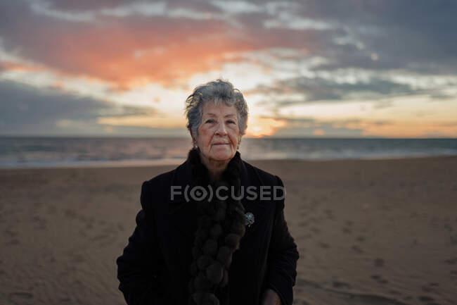 Feminino sênior em roupas casuais quentes admirando o pôr do sol sobre o mar enquanto descansa sozinho na praia — Fotografia de Stock
