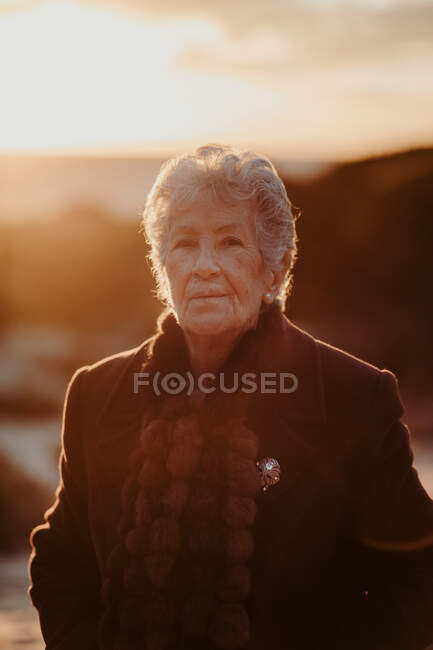 Elderly female tourist with gray hair in warm casual outfit looking at camera while relaxing on sandy beach against cloudy evening sky — Stock Photo