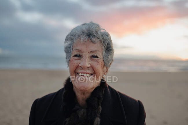 Femme âgée en vêtements décontractés chauds admirant coucher de soleil sur la mer tout en se reposant seul sur la plage de sable — Photo de stock