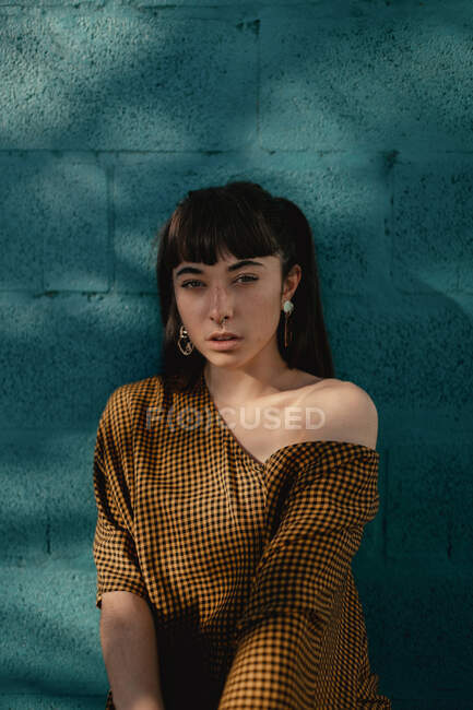 Стильна молода етнічна жінка з довгим темним волоссям в модному платті, що стоїть на зеленій стіні на вулиці і дивиться на камеру вдумливо — стокове фото
