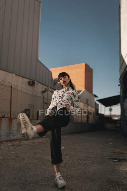 Full length of stylish self assured young ethnic female millennial in trendy outfit looking at camera while standing in urban area against cloudless blue sky with raised leg — Stock Photo