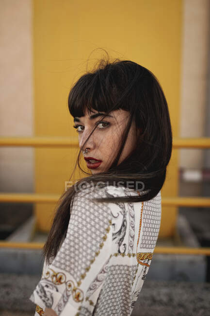 Side view young self esteem ethnic female with long dark hair in stylish outfit standing near metal railing near modern building and looking at camera — Stock Photo