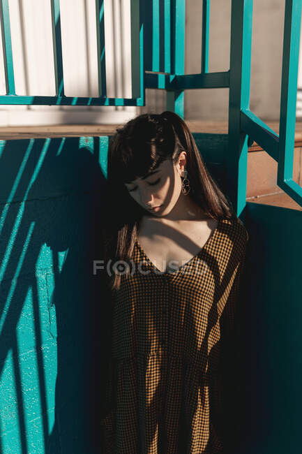Стильная молодая этническая женщина с длинными темными волосами в модном платье, стоящая напротив зеленой стены на улице и задумчиво смотрящая вниз — стоковое фото