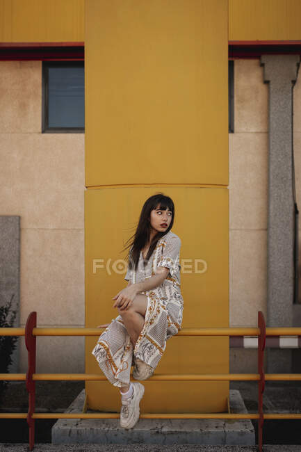 Полное тело молодой самооценки этнической женщины с длинными темными волосами в стильном наряде сидит на металлических перилах рядом с современным зданием и смотрит в сторону — стоковое фото