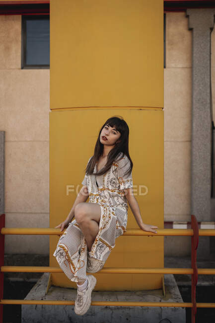 Cuerpo completo de joven autoestima étnica femenina con el pelo largo y oscuro en traje elegante sentado en barandilla de metal cerca de edificio moderno y mirando a la cámara - foto de stock