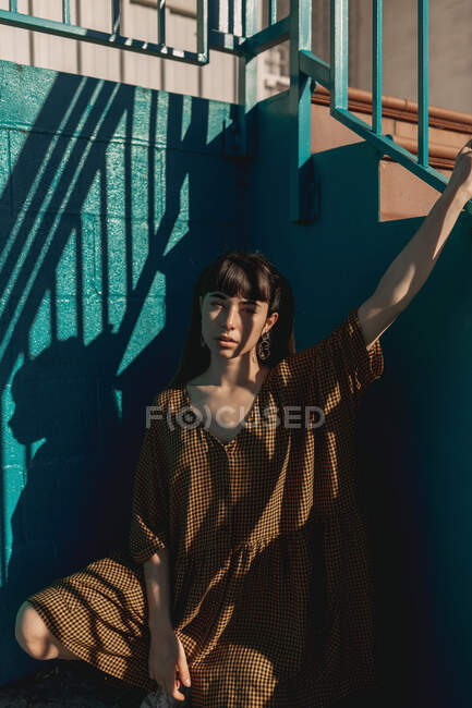 Alluring young ethnic lady with long dark hair in stylish dress sitting on haunches and holding metal railing of staircase on city street — Stock Photo