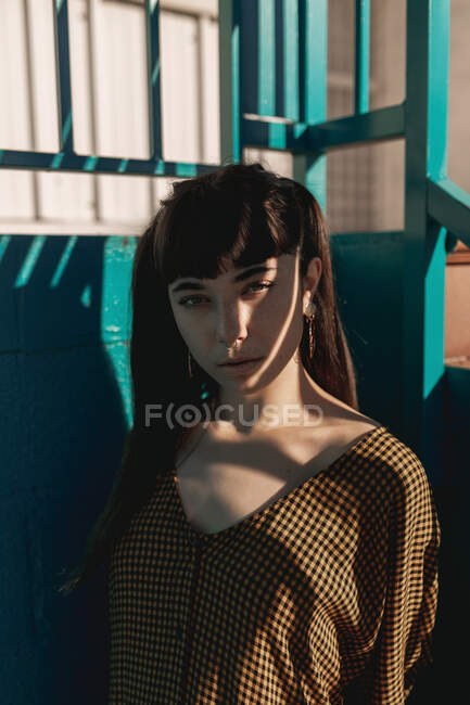 Stilvolle junge ethnische Frau mit langen dunklen Haaren im trendigen Kleid steht an einer grünen Mauer auf der Straße und blickt nachdenklich in die Kamera — Stockfoto