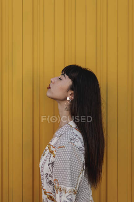 Вид сбоку на стильную молодую этническую женщину с длинными темными волосами в модном платье, стоящую напротив желтой стены на улице и задумчиво закрытые глаза — стоковое фото