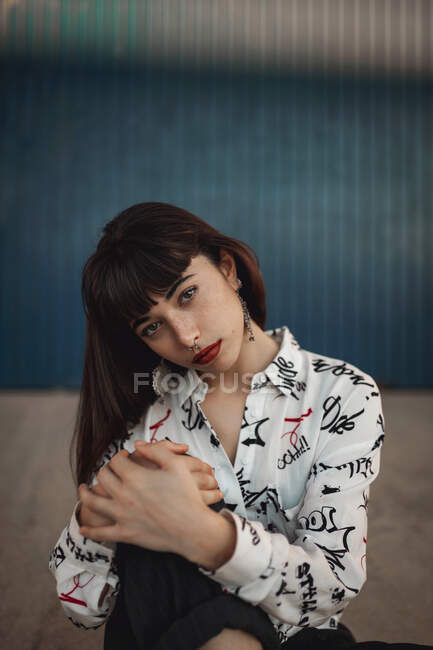 Dreamy young ethnic female millennial with long dark hair in casual clothes embracing knee and looking at camera on street — Stock Photo