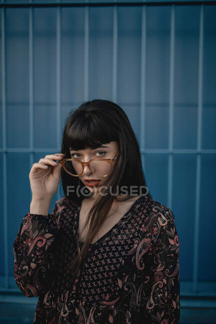 Stylish young ethnic female with long dark hair in trendy dress standing against blue wall on street and looking at camera thoughtfully — Stock Photo