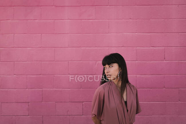 Side view of stylish young ethnic female with long dark hair in trendy dress standing against pink wall on street and looking away thoughtfully — Stock Photo