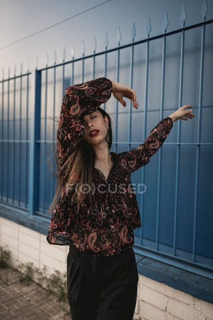 Elegante joven étnica femenina con el pelo largo y oscuro en vestido de moda de pie contra la pared azul en la calle y los ojos cerrados pensativamente - foto de stock