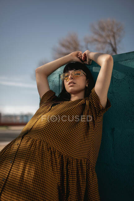 Baixo ângulo de auto-seguro jovem étnico feminino em vestido elegante e óculos de sol apoiados na parede com braços levantados e olhando para a câmera na rua — Fotografia de Stock