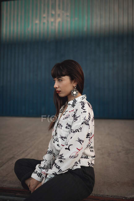 Dreamy young ethnic female millennial with long dark hair in casual clothes sitting on floor and looking away on street — Stock Photo