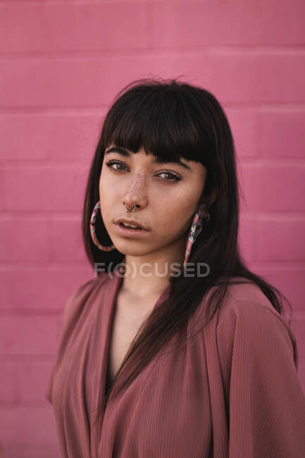Jeune femme ethnique élégante avec de longs cheveux foncés en robe tendance debout contre un mur rose dans la rue et regardant la caméra avec attention — Photo de stock