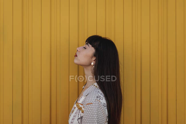 Side view of stylish young ethnic female with long dark hair in trendy dress standing against yellow wall on street and closed eyes thoughtfully — Stock Photo