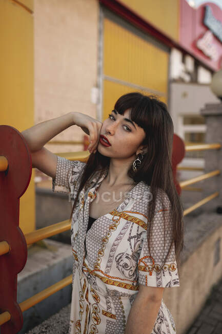 Young self esteem ethnic female with long dark hair in stylish outfit leaning on metal railing near modern building and looking at camera — Stock Photo