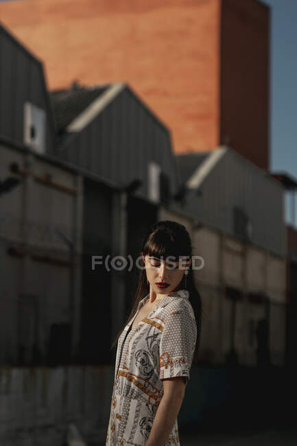 Vista lateral de la joven mujer étnica pensativa con el pelo largo y oscuro en vestido casual de pie en la calle cerca de casas viejas y mirando hacia abajo en el día soleado - foto de stock