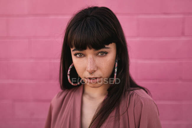 Jeune femme ethnique élégante avec de longs cheveux foncés en robe tendance debout contre un mur rose dans la rue et regardant la caméra avec attention — Photo de stock