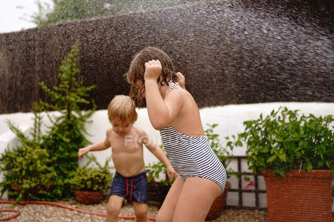 Вид сбоку веселый мальчик без рубашки льет воду из шланга на сестру в купальнике, играя вместе во дворе в летний день — стоковое фото