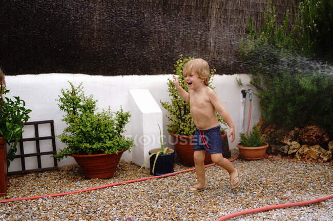 Вид збоку милий маленький хлопчик у купальнику ходить і поливає зелену галявину з шланга під час літніх канікул у сільській місцевості — стокове фото