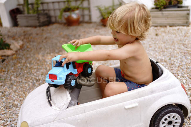 Vista lateral de lindo niño alegre con pelo rubio montar coche de juguete mientras se encuentra en el patio en el día de verano soleado - foto de stock