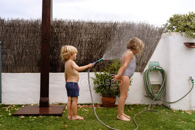 Side view of cheerful shirtless little boy pouring water from hose on sister in swimsuit while playing together in yard on summer day — Stock Photo