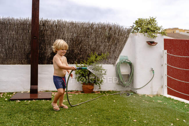 Vista lateral do menino bonito em maiô andando e regando gramado verde da mangueira durante as férias de verão no campo — Fotografia de Stock