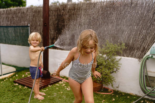 Alegre sin camisa niño vertiendo agua de la manguera en la hermana en traje de baño mientras juegan juntos en el patio en el día de verano - foto de stock