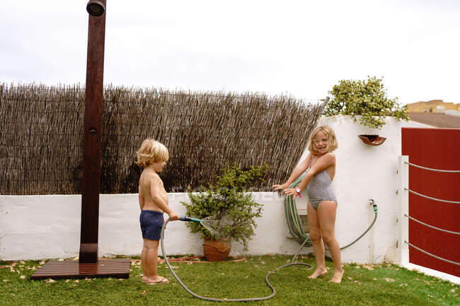 Seitenansicht des fröhlichen, hemdlosen kleinen Jungen, der an einem Sommertag im Hof zusammen mit seiner Schwester im Badeanzug Wasser aus dem Schlauch gießt — Stockfoto