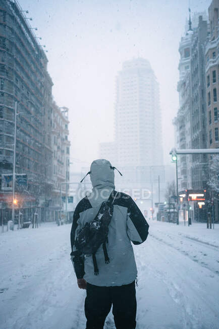 На задньому плані зображений анонімний чоловік у зовнішньому одязі з теплим одягом, що йде по вулиці з будинками в сніжну зиму в Мадриді. — стокове фото