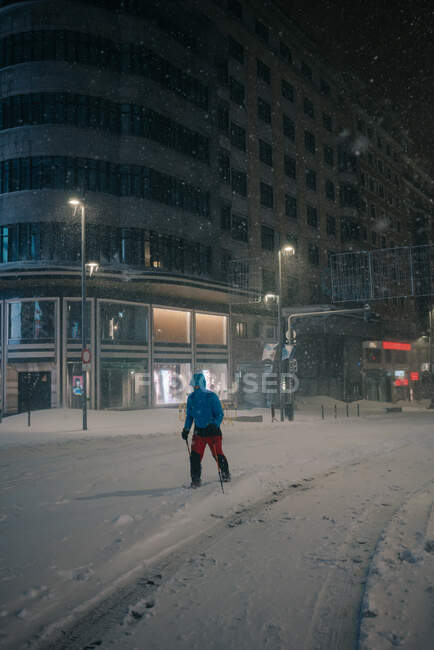 Atleta masculino anônimo em sportswear esqui na estrada nevada contra a construção à noite durante a queda de neve em Madrid Espanha — Fotografia de Stock