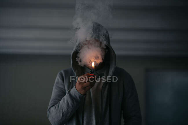 Unrecognizable male in hooded jacket smoking cigarette while standing in dark room with burning lighter in hand — Stock Photo
