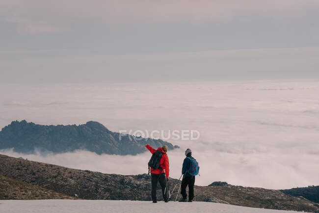 Вид ззаду анонімних дослідників у теплому верхньому одязі з рюкзаками та паличками, що стоять на засніженому піку гори, прихованому під товстими хмарами — стокове фото
