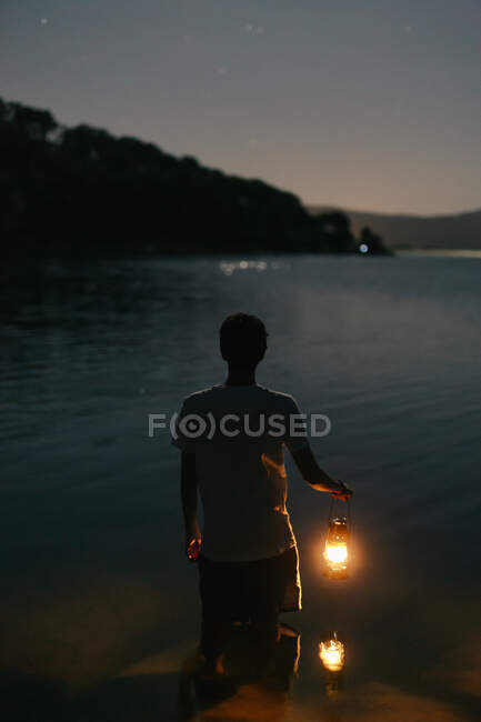 Back view of faceless male tourist with shiny lantern contemplating rippled river against mount at dusk — Stock Photo