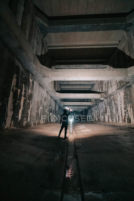 Back view of anonymous person with glowing flashlight in hand standing in abandoned tunnel with shabby walls — Stock Photo