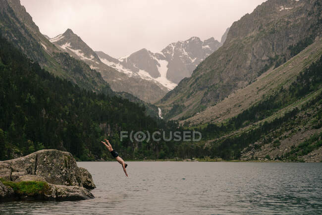 Vista lateral de un viajero irreconocible sin camisa saltando al agua del lago rodeado de enormes montañas rocosas en un día nublado - foto de stock