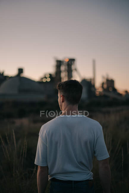 Задний вид анонимного мужчины на лугу против электростанции под серым небом в вечернем поле — стоковое фото