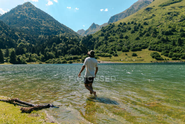 Back view of anonymous male traveler admiring high mountains while standing in pure ocean water during summer trip — Stock Photo