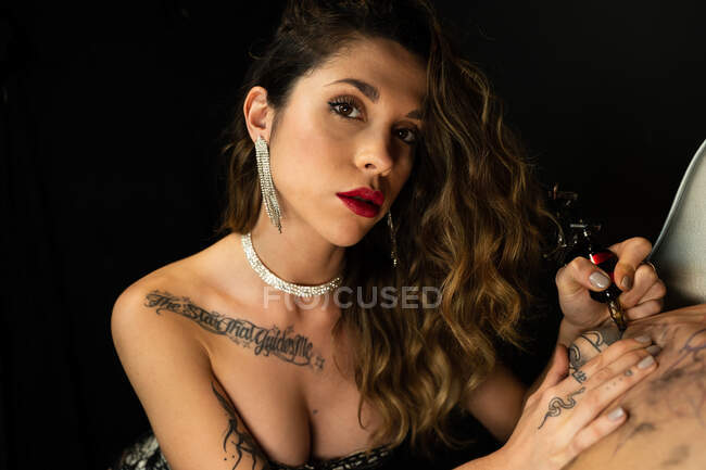 Sexy female tattoo master with machine making tattoo on arm of male customer while looking at camera in dark salon — Stock Photo