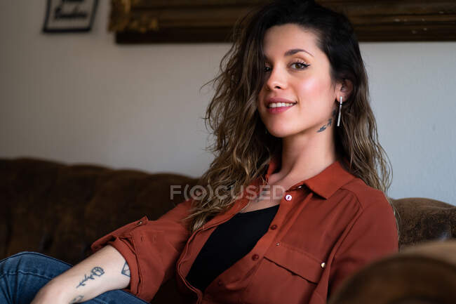 Conteúdo feminino em desgaste casual e com tatuagens sentadas no sofá e olhando para a câmera — Fotografia de Stock