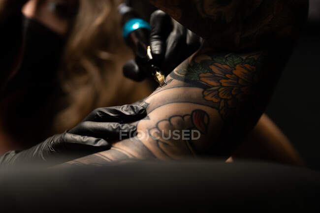 Mestre de tatuagem feminina em máscara e com máquina fazendo tatuagem no braço do cliente masculino no salão escuro — Fotografia de Stock