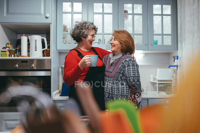 Zufriedene homosexuelle reife Partnerinnen mit einer Tasse Kaffee, die sich zu Hause umarmen und anschauen — Stockfoto