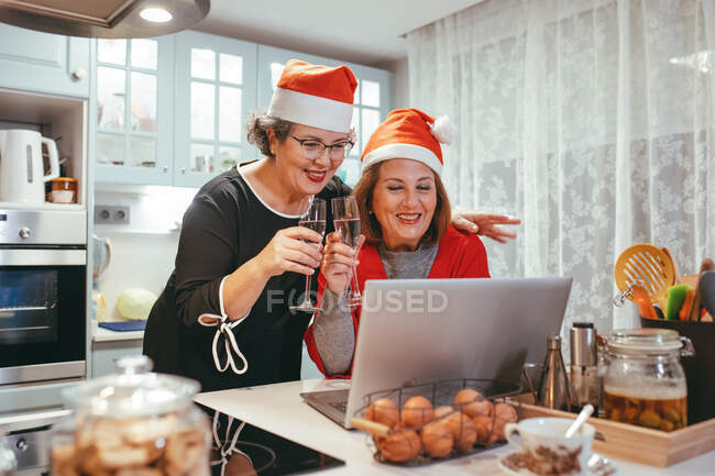 Mujeres maduras homosexuales sinceras en sombreros de Santa con champán viendo netbook con chat de vídeo durante las vacaciones de Año Nuevo en la cocina - foto de stock