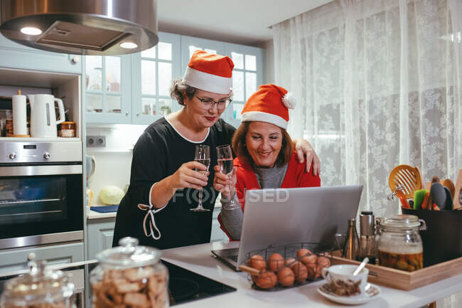 Donne mature omosessuali sincere a Babbo Natale cappelli con champagne guardando netbook con video chat durante le vacanze di Capodanno in cucina — Foto stock