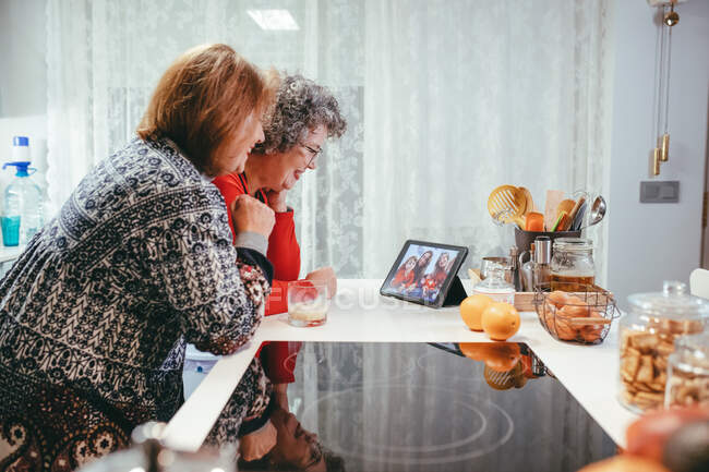 Visão lateral de mulheres idosas homossexuais sorridentes assistindo tablet com crianças e mães felizes durante o bate-papo por vídeo em casa — Fotografia de Stock