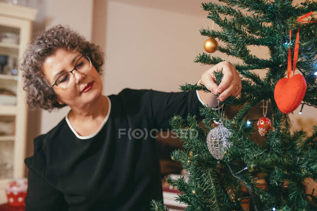 Focused middle aged female in eyeglasses with tilted head decorating fir tree before New Year holiday in house — Stock Photo